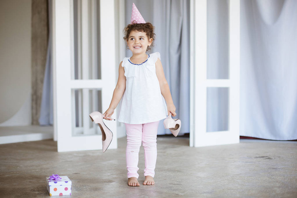 Radosna dziewczynka, ciemne kręcone włosy w urodziny cap, szczęśliwie patrząc na bok trzyma buty na obcasach w rękach pudełko w pobliżu na piętrze w domu - Zdjęcie, obraz