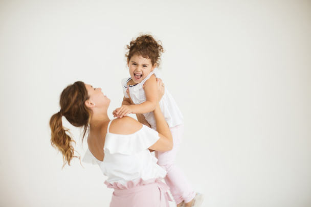 Молодая мама держит в руках маленькую милую дочь, которая радостно смотрит в камеру, проводя время вместе на белом фоне
 - Фото, изображение