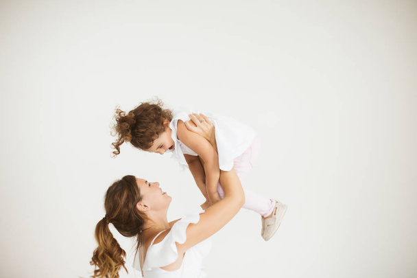Young beautiful mother happily lifting little cute daughter up spending time together over white background - Zdjęcie, obraz