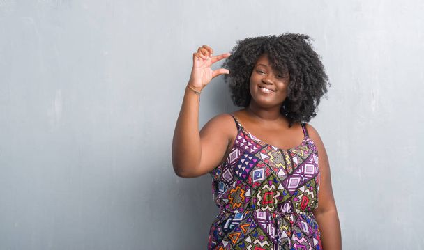 Mladá africká americká žena přes zeď šedá grunge nosí barevné šaty s úsměvem a jistý ukázal rukou dělá znamení velikost prsty při pohledu a fotoaparát. Opatření koncepce. - Fotografie, Obrázek