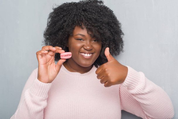 Νεαρός αφρικανική αμερικανική γυναίκα πάνω από την γκρίζα grunge τοίχο τρώει ροζ macaron ευχαριστημένοι με μεγάλο χαμόγελο κάνει εντάξει σημάδι, αντίχειρα επάνω με τα δάχτυλα, καλό σημάδι - Φωτογραφία, εικόνα