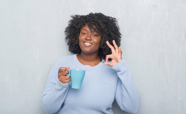 Jovem afro-americana sobre parede grunge cinza bebendo uma xícara de café fazendo sinal ok com os dedos, excelente símbolo
 - Foto, Imagem