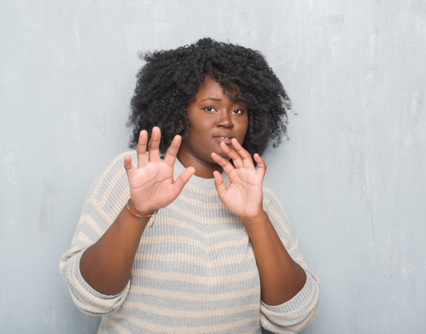 Молода афроамериканка плюс розмір жінка над сірою гранжевою стіною, одягнена в светр, боїться і наляканий виразом страху зупинити жест руками, кричачи в шоці. концепція паніки
. - Фото, зображення