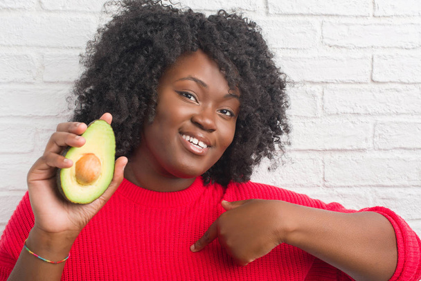 Fiatal afrikai-amerikai nő túltáplálkozás fehér tégla fal avokádó mutatóujjal meglepetés arc magát - Fotó, kép