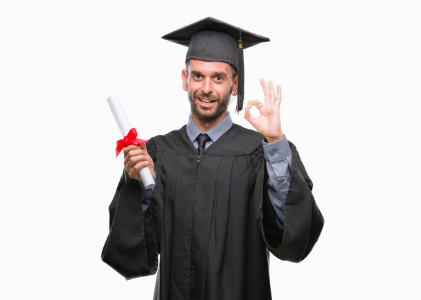 junge gut aussehende graduierte Mann mit Abschluss über isolierten Hintergrund tun ok Zeichen mit Fingern, ausgezeichnetes Symbol - Foto, Bild