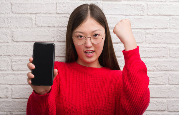 Jonge Chinese vrouw over bakstenen muur weergegeven: smartphone boos en gefrustreerd schreeuwen met woede, gek en schreeuwen met opgeheven hand, woede concept - Foto, afbeelding