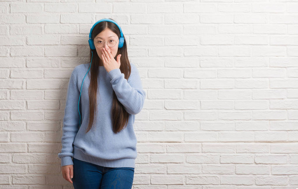 Giovane donna cinese oltre muro di mattoni indossando cuffie coprire la bocca con la mano scioccata dalla vergogna per errore, espressione di paura, paura in silenzio, concetto segreto
 - Foto, immagini