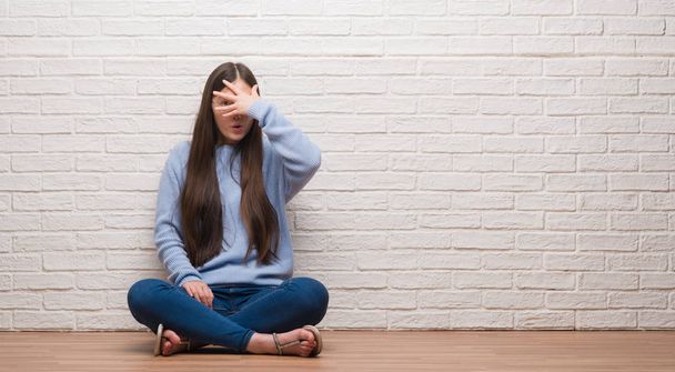 Young Chinese woman sitting on the floor over brick wall peeking in shock covering face and eyes with hand, looking through fingers with embarrassed expression. - Foto, Imagen