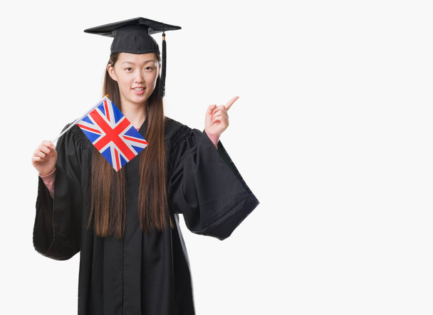 Joven mujer china con uniforme de graduado sosteniendo bandera del Reino Unido muy feliz señalando con la mano y el dedo a un lado
 - Foto, Imagen