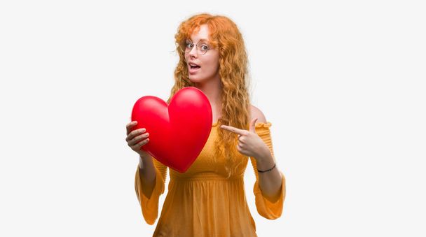 Nuori punapää nainen rakastunut tilalla punainen sydän erittäin onnellinen osoittaa kädellä ja sormella
 - Valokuva, kuva