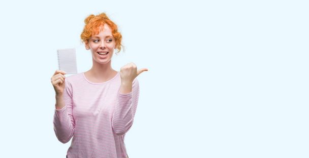 Jonge roodharige vrouw met lege laptop aan te wijzen en te tonen met de duim aan de kant met een blij gezicht lachend - Foto, afbeelding