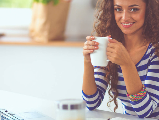 Smiling young woman with coffee cup and laptop in the kitchen at home - Foto, immagini