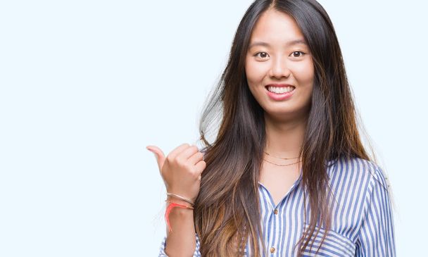 Young asian woman over isolated background smiling with happy face looking and pointing to the side with thumb up. - 写真・画像