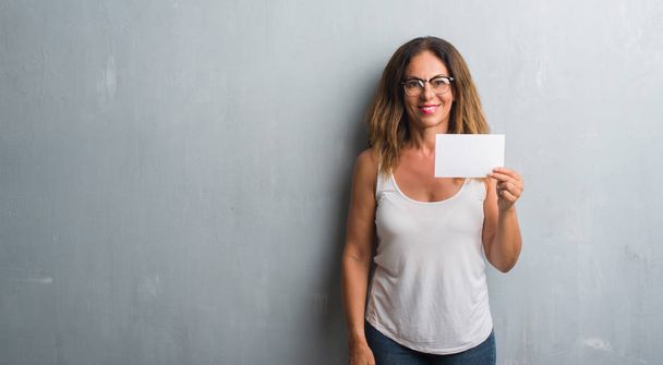 Латиноамериканка средних лет, стоящая над серой гранж-стеной, держит чистую карточку со счастливым лицом, стоя и улыбаясь с уверенной улыбкой, показывая зубы.
 - Фото, изображение