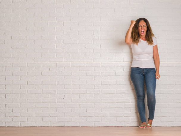 Mujer hispana de mediana edad de pie sobre la pared de ladrillo blanco molesto y frustrado gritando con ira, loco y gritando con la mano levantada, concepto de ira
 - Foto, imagen