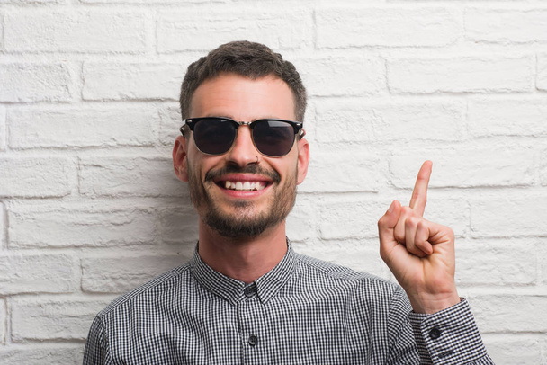 Young adult man wearing sunglasses standing over white brick wall surprised with an idea or question pointing finger with happy face, number one - Photo, Image