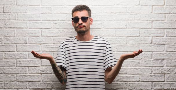 Молодой взрослый мужчина в солнечных очках, стоящий над белой кирпичной стеной, невежественное и запутанное выражение лица с поднятыми руками. Концепция сомнений
. - Фото, изображение