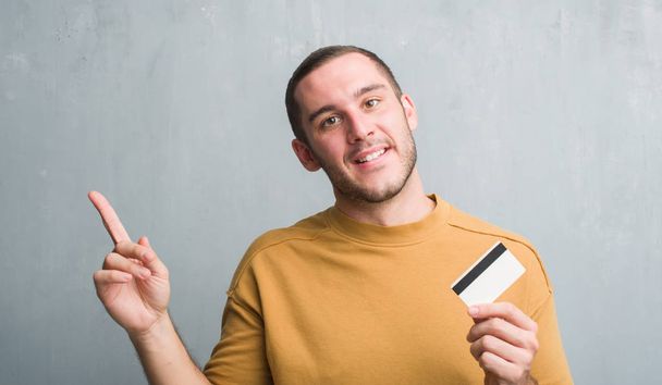 Kaukázusi fiatalember gazdaság hitelkártya nagyon boldog, mutat, a kéz és az ujjak oldalán szürke grunge falon - Fotó, kép