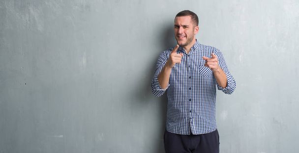 Homem caucasiano jovem sobre parede grunge cinza apontando os dedos para a câmera com rosto feliz e engraçado. Boa energia e vibrações
. - Foto, Imagem