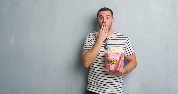 Nuori valkoihoinen mies harmaan murojen päällä syömässä popcornpeitteen suuta käsi järkyttyneenä erehdyksestä, pelon ilmauksesta, pelosta hiljaisuudessa, salaisesta käsitteestä
 - Valokuva, kuva