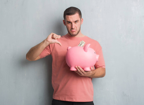 Young caucasian man over grey grunge wall holding piggy bank with angry face, negative sign showing dislike with thumbs down, rejection concept - Photo, Image