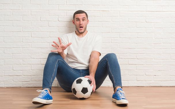 Молодий білий чоловік сидить над білою цегляною стіною, тримаючи футбольний м'яч, наляканий шоком з несподіваним обличчям, боїться і схвильований виразом страху
 - Фото, зображення