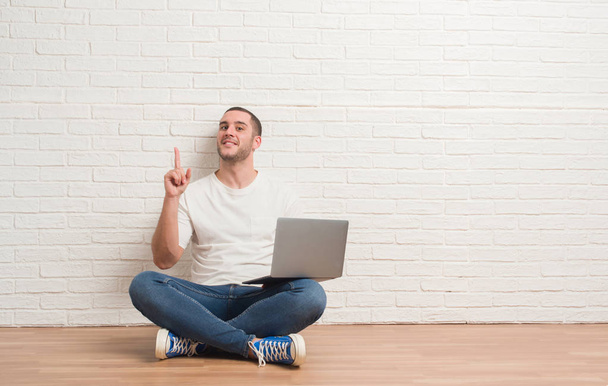 Młody człowiek kaukaski siedzi nad białym murem przy użyciu komputera przenośnego zaskoczony z pomysłu lub pytanie palcem wskazującym z szczęśliwy twarz, numer jeden - Zdjęcie, obraz