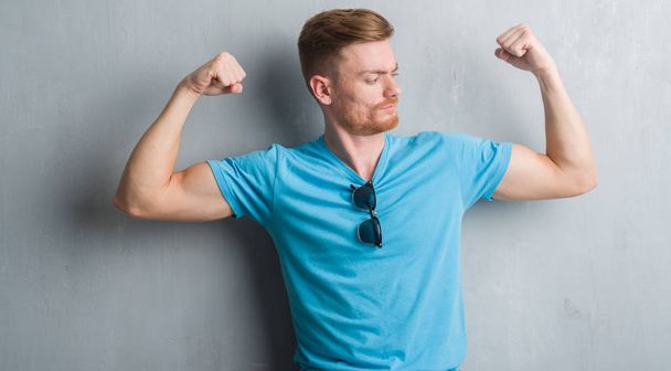 Jonge roodharige man over grijs grunge muur casual outfit weergegeven: armen spieren glimlachend trots dragen. Fitness concept. - Foto, afbeelding