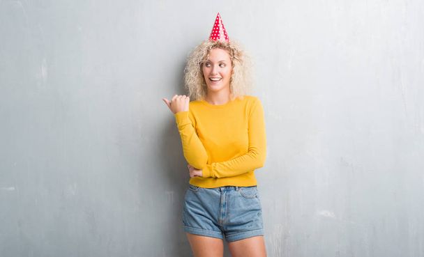 Jeune femme blonde sur le mur gris grunge portant un chapeau d'anniversaire souriant avec un visage heureux regardant et pointant vers le côté avec le pouce vers le haut
. - Photo, image