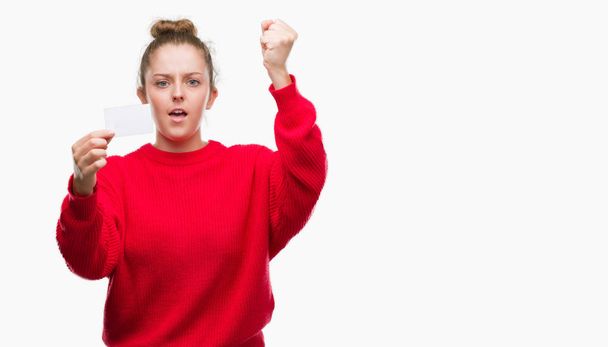 Jonge blonde vrouw met reclame kaart boos en gefrustreerd schreeuwen met woede, gek en schreeuwen met opgeheven hand, woede concept - Foto, afbeelding