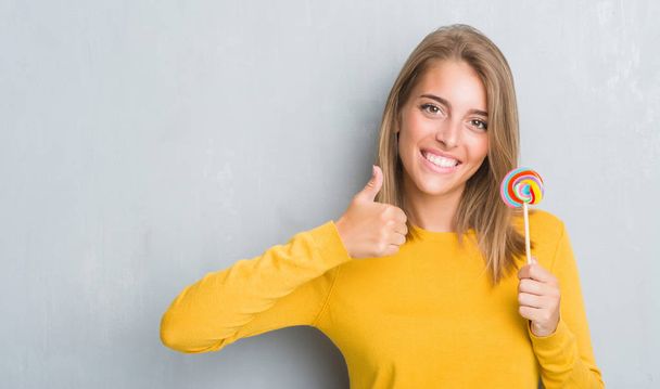 Beautiful young woman over grunge grey wall eating lollipop candy happy with big smile doing ok sign, thumb up with fingers, excellent sign - Photo, Image