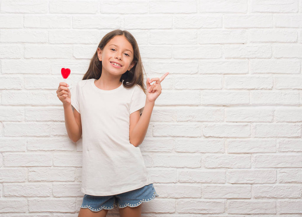 Giovane bambino ispanico sopra muro di mattoni bianchi mangiare caramelle cuore rosso lecca-lecca molto felice che punta con mano e dito di lato
 - Foto, immagini