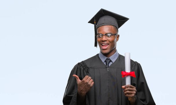 Young graduated african american man holding degree over isolated background pointing and showing with thumb up to the side with happy face smiling - Foto, immagini