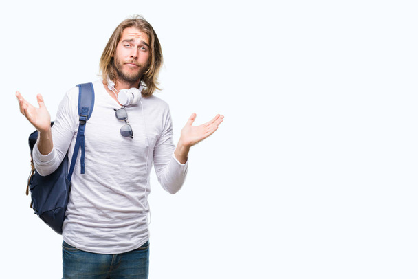 Genç yakışıklı turist adam uzun saçlı izole arka plan clueless ve şaşkın bir ifadeyle kollar ve eller kaldırdı üzerinde sırt çantası giyiyor. Şüphe kavramı. - Fotoğraf, Görsel