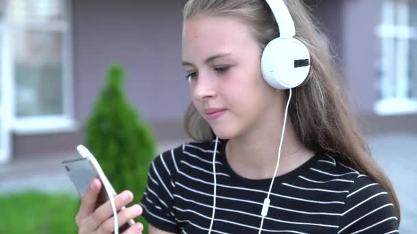 Young teenage woman with light brown hair and white headphones grinning while looking at her smart phone - Felvétel, videó