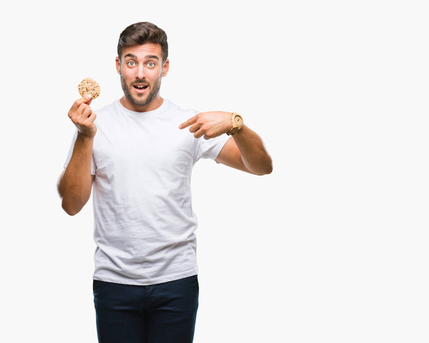 Young handsome man eating chocolate chips cookie over isolated background with surprise face pointing finger to himself - Foto, Imagen