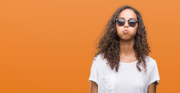 Jeune femme hispanique portant des lunettes de soleil gonflant les joues avec un visage drôle. Bouche gonflée avec de l'air, expression folle
. - Photo, image