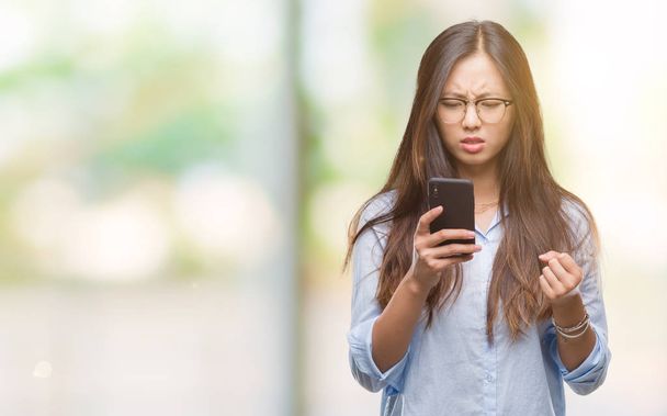 Jonge Aziatische vrouw texting gebruik van smartphone over geïsoleerde achtergrond boos en gefrustreerd schreeuwen met woede, gek en schreeuwen met opgeheven hand, woede concept - Foto, afbeelding