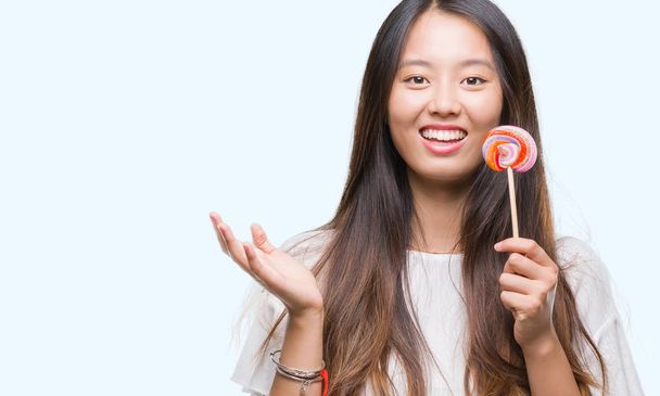 Genç Asyalı kadın üzerinde lolipop şeker yemek büyük gülümseme ile çığlık zaferini kutluyor ve ellerini kaldırdı arka plan çok mutlu ve heyecanlı, kazanan ifade izole - Fotoğraf, Görsel