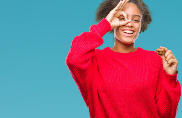 Jeune femme afro-américaine mangeant du macaron au chocolat sur fond isolé avec un visage heureux souriant faisant signe ok avec la main sur les yeux regardant à travers les doigts
 - Photo, image