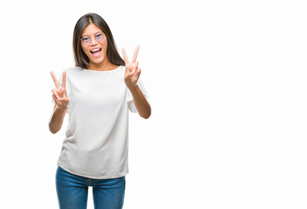 Młoda kobieta azjatyckich w okularach na białym tle uśmiechnięty z szczęśliwy twarz mrugając aparat robi znak zwycięstwa. Numer dwa. - Zdjęcie, obraz