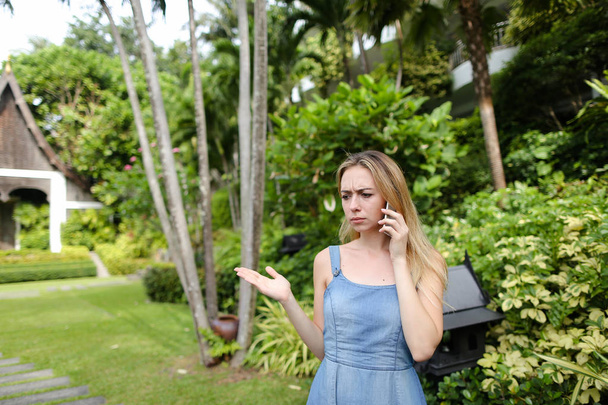 Mujer joven hablando por teléfono inteligente en el jardín cerca de los árboles, con pantalones vaqueros vestido
. - Foto, imagen