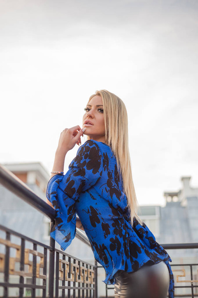 blonde woman in blue blouse and black leather leggings posing near railing on balcony  - 写真・画像