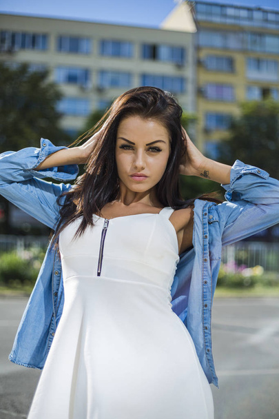 attractive pretty woman in white dress with jeans jacket looking at camera while standing on street  - Fotografie, Obrázek