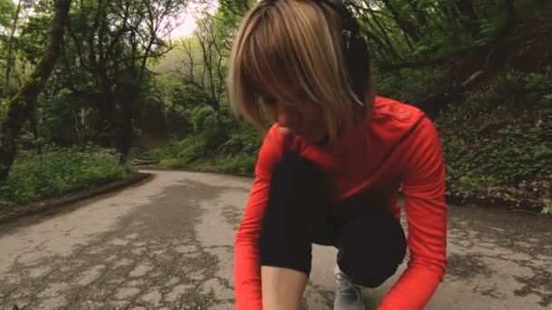 Młoda dziewczyna Kaukaska siedzi wiąże jej sznurowadła przed jogging na leśnej drodze na Kaukazie. Szeroki kąt - Materiał filmowy, wideo