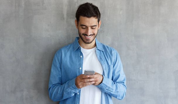 Young smiling man wearing blue shirt, standing against gray textured wall with copy space, looking at screen of smartphone - Zdjęcie, obraz