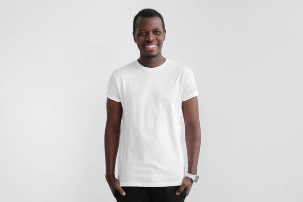 Young smiling african man with dark skin standing with hands in pockets, wearing blank white t shirt with copy space for your logo or text, isolated on gray background - 写真・画像