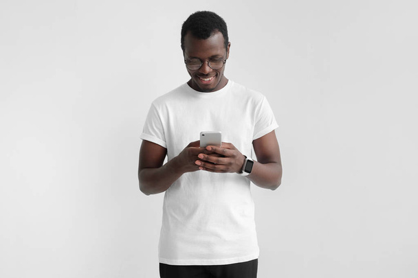 Daylight portrait of young african american man isolated on gray background wearing white t-shirt standing in front of camera, looking attentively with smile at screen of smartphone he is holding - Foto, imagen