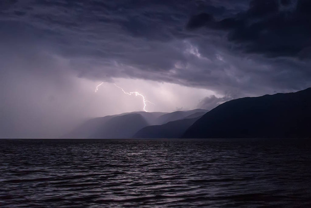 Amazing view with a flash of lightning in dark blue clouds during a thunderstorm over a lake in the mountains - Фото, изображение
