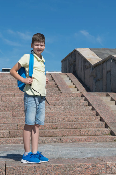 the schoolboy is standing on the stairs with a blue backpack for school - Foto, Imagem